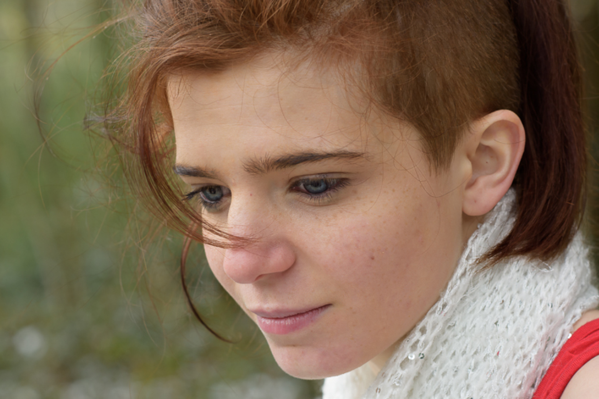 Portrait rapproché d’une femme en extérieur, regard vers le bas, cheveux légèrement au vent, lumière naturelle douce et arrière-plan flou