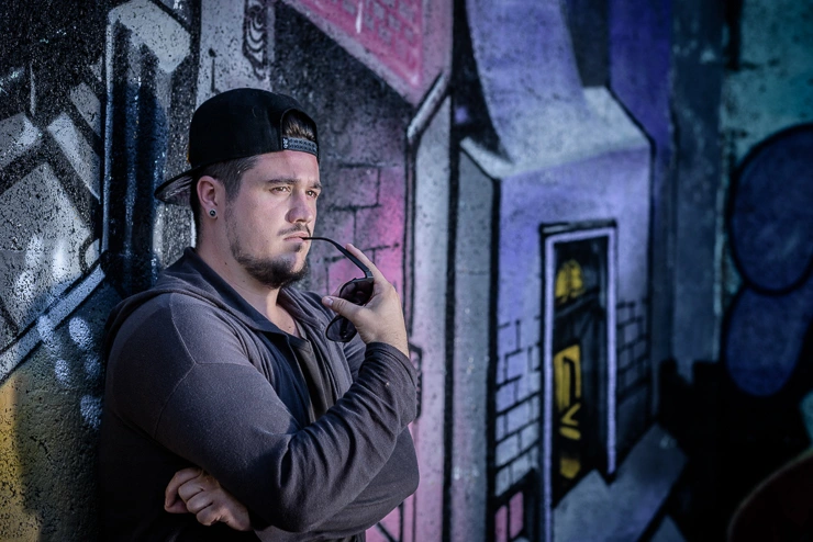 Homme en portrait appuyé contre un mur avec graffiti, regard concentré, tenant des lunettes de soleil, éclairage naturel contrasté