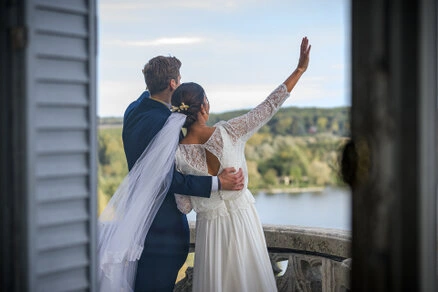 Couple de mariés de dos regardant un paysage et levant la main depuis une terrasse lors d’un mariage