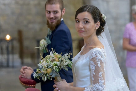 La mariée tenant son bouquet regarde vers l’objectif pendant la cérémonie, avec le marié en arrière-plan