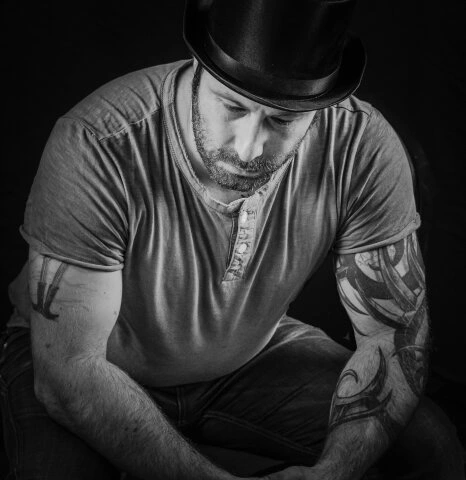 Portrait en noir et blanc d’un homme assis portant un chapeau, regard baissé, lumière contrastée et fond sombre