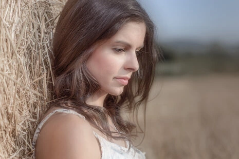 Portrait d’une femme en extérieur appuyée contre une botte de paille, regard vers le bas, lumière douce et arrière-plan flou