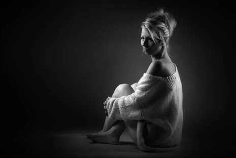 Portrait d’une femme assise en noir et blanc, éclairée par une lumière douce créant des contrastes subtils et une ambiance intimiste