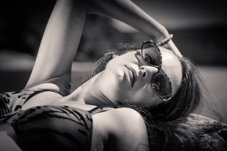 Portrait en noir et blanc d’une femme allongée en extérieur, portant des lunettes de soleil, pose détendue et lumière contrastée