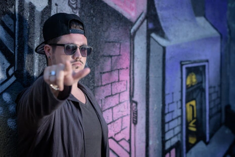 Portrait d’un homme en environnement urbain avec lunettes et casquette, faisant un geste vers l’objectif devant un mur graffiti