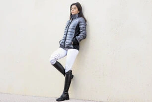 Femme adossée à un mur clair, portant une doudoune, un pantalon ajusté et des bottes hautes, avec une posture détendue et un regard dirigé sur le côté