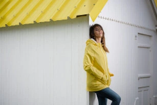 Femme en veste jaune appuyée contre une structure aux lignes graphiques, dans une mise en scène de style mode contemporain