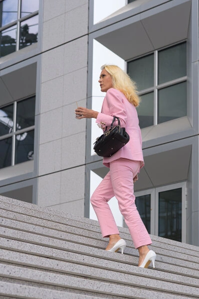 Femme en tailleur rose et escarpins blancs montant un escalier en extérieur, tenant un sac à main, avec un décor architectural moderne en arrière-plan