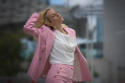 Femme en tailleur rose avec un top blanc, la tête légèrement en arrière et la main dans les cheveux, dans un moment de mouvement capté en extérieur