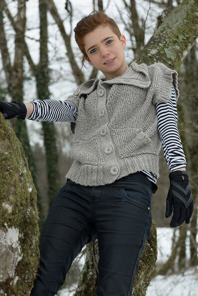 Femme appuyée contre un arbre en extérieur, portant un gilet en maille et des gants, dans un environnement naturel avec une ambiance hivernale