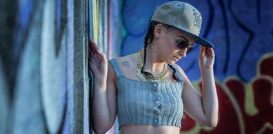 Femme portant une casquette, des lunettes de soleil et un débardeur, adossée à un mur avec graffiti, avec une posture naturelle et un regard dirigé vers le bas
