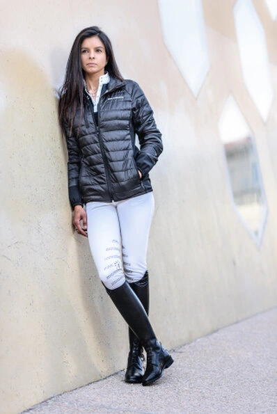 Femme adossée à un mur clair, portant une doudoune noire, un pantalon ajusté et des bottes hautes, avec une posture inclinée et un regard dirigé sur le côté