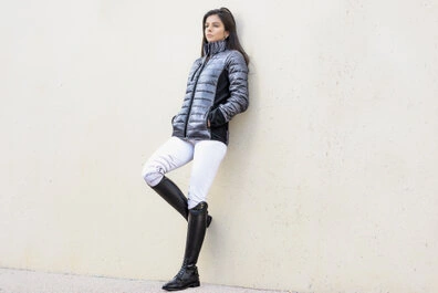 Femme adossée à un mur clair, portant une doudoune, un pantalon ajusté et des bottes hautes, avec une posture détendue et un regard dirigé sur le côté