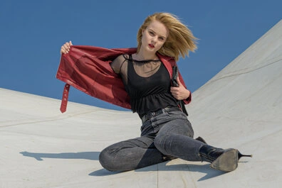 Femme en veste rouge ouverte et jean sombre, assise sur une surface claire avec une posture dynamique, regardant vers l’objectif sous un ciel bleu