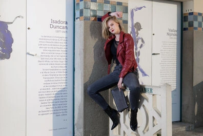 Femme en veste rouge et jean assise sur un élément en béton, avec une posture posée et un mur comportant des éléments graphiques et du texte en arrière-plan