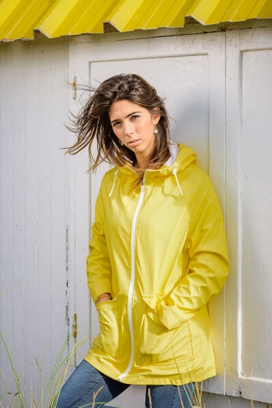 Femme portant une veste jaune et un jean, adossée à une structure en bois blanc avec un élément jaune au-dessus, regardant vers l’objectif