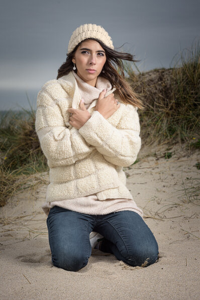 Femme portant un pull en maille, un bonnet et une écharpe, à genoux sur du sable avec des herbes en arrière-plan, dans une ambiance froide et ventée