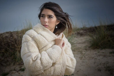 Femme portant un pull en maille épaisse sur une dune avec des herbes en arrière-plan, tenant le col près du cou avec une expression calme et un regard dirigé sur le côté