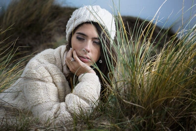 Femme allongée dans les herbes sur une dune, portant un pull en maille et un bonnet, la tête appuyée sur la main avec un regard dirigé vers le bas